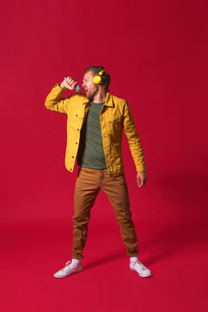 Singing young man enjoying his favorite song holding microphone and wireless headphones wearing casual jacket isolated on red background. Man sing while recording his voice. A star is born.の写真素材