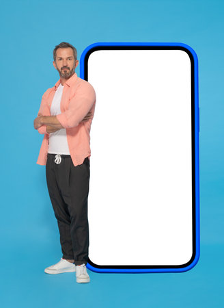 Posing with arms crossed in front big smartphone with white screen in blue case middle aged grey haired man wearing peach shirt and jogger pants isolated on blue background. Mobile app advertising.の写真素材