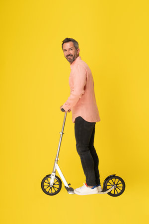 Middle aged grey haired man ride scooter standing sideways looking at camera wearing peach shirt and black jeans isolated on yellow background. Fit masculine handsome mature businessman on the go.の写真素材