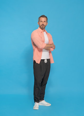 Trendy middle aged grey haired man posing with arms folded looking at camera wearing pink shirt and black jogger pants isolated on yellow background. Mature fit man, health lifestyle concept.の写真素材