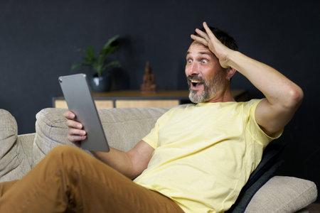 Middle aged handsome man use digital tablet to communicate via video call or reading news with shock facial expression sitting on the sofa in home interior wearing casual. Freelancer, work from home.の写真素材