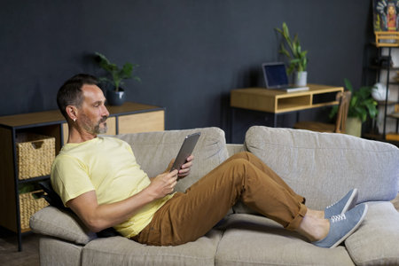 Working offline or watching movie laying on the couch middle aged freelancer man use digital tablet at cozy home wearing casual. Relaxed freelancer man laying on sofa making business uses gadget.の写真素材
