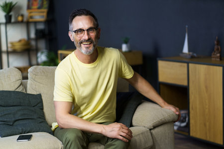 Cheerful grey haired middle aged man sitting on the sofa wearing glasses looking at camera wearing casual. Thoughtful freelancer mature man working from home.の写真素材