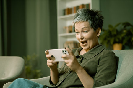 Woman at home is fully engrossed in her phone game, with a big smile on her face as she plays. Her happiness is evident as she focuses on achieving her goals in the game. High quality photoの写真素材