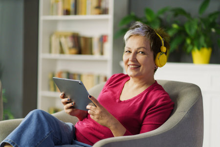 Mature woman smiles on a sofa, holding tablet PC and listening to music on headphones. She enjoys leisure, technology and music, feeling tranquil and blissful. High quality photoの写真素材