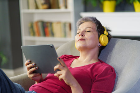 Mature woman captured on sofa home holding tablet PC and wearing headphones while listening music with her eyes closed. Woman enjoys her leisure time while indulging in pleasures of modern technology.の写真素材