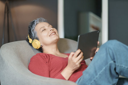 Mature woman with grey hair is depicted listening to music while sitting on sofa in her home. She has her eyes closed and is holding a tablet in her hands. High quality photoの写真素材