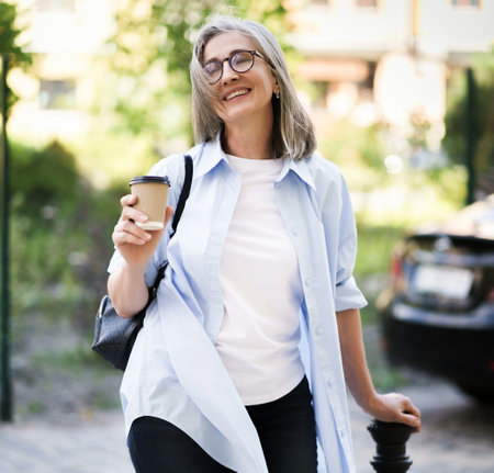 Happy mature woman enjoying a cup of coffee on a summer day while sitting outdoors on a city street. She appears content and relaxed, savoring the simple pleasures of life. High quality photoの写真素材