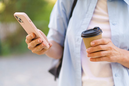 close-up photo focuses on a womans hands holding a mobile phone and a coffee paper cup. The image depicts modern lifestyle and the widespread use of technology in daily routines. . High quality photoの写真素材
