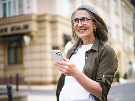 Joyful senior woman with grey hair and mobile phone in hand, enjoying her time in beautiful European city. Image showcases active and modern urban lifestyle of the elderly generation. High quality photoの写真素材
