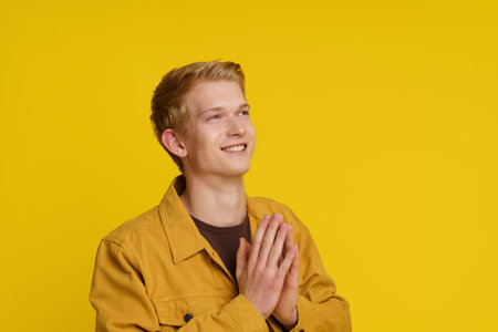 Happiness Gratitude Faith Concept. Smiling Young European Man Joined His Palms in a Gesture of Gratitude on a Yellow Background. High quality photoの写真素材