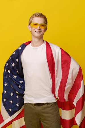 Super American Hero Concept. Youth Smiles in a White T-shirt and the American Flag is isolated on a yellow background. High quality photoの写真素材
