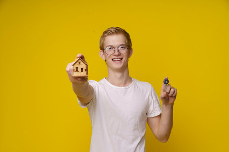 Young man holds a model of a house and a key, symbolizing his independence and entry into the housing market. This image represents the concept of affordable real estate for the youth and the importance of financial planning and property ownership. High quality photoの写真素材