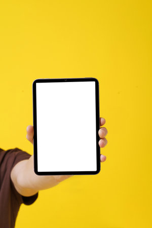 close-up image of a mans hand holding a blank screen Tablet PC against a yellow background,...