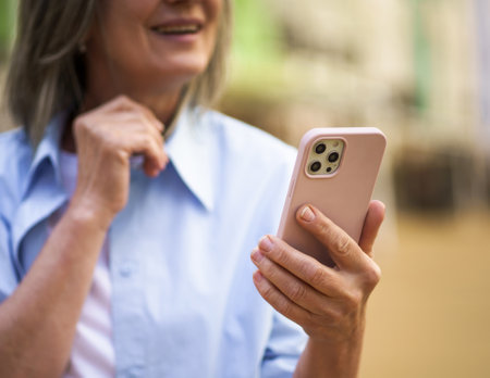 Close-up photo captures hands of elderly woman holding smartphone in old city. The senior womans smile and positive expression convey sense of happiness and connection through technology. High quality photoの写真素材