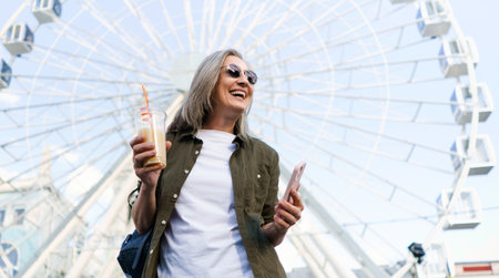 Cheerful senior woman enjoys a sunny day in an old European city, holding her phone and a cup of juice in her hands with a big smile on her face, embodying the concept of elderly happiness. High quality photoの写真素材