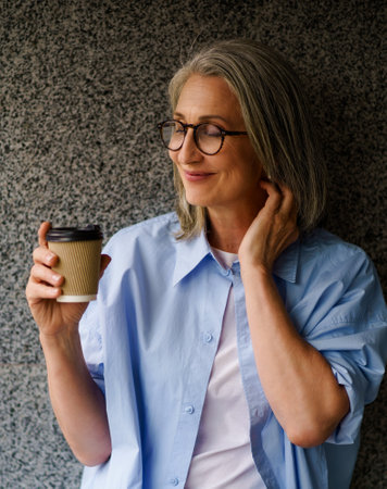 Joyful and stylish senior woman with coffee cup in her hand, standing near wall in urban city setting. She radiates happiness and relaxation, making it the perfect image to illustrate carefree and content lifestyle. High quality photoの写真素材