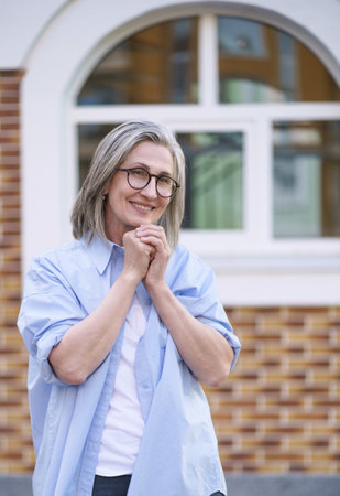 Older woman in a city setting, smiling with her hands clasped together in front of her, conveying a sense of happiness and contentment. High quality photoの写真素材