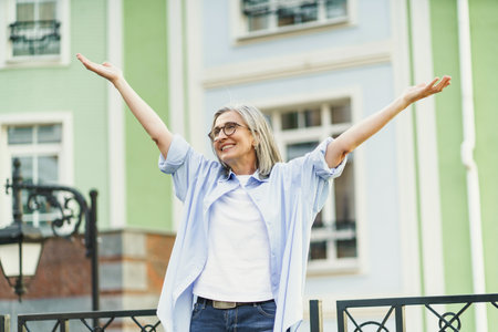 Mature woman standing in old city, smiling and raising her hands to sky, expressing happiness and positive emotions. Perfect illustration of happiness in everyday life and senior living. High quality photoの写真素材