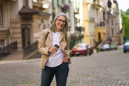 Mature woman with cheerful smile holding phone while exploring old European city. Photo captures concept of elderly happiness and technology use for communication and travel purposes. High quality photoの写真素材