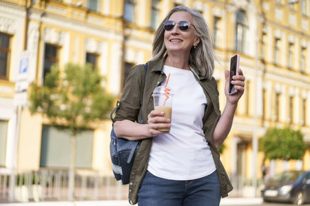 Biohacking Concept. A Mature European Woman with Gray Hair wearing Sunglasses Walks around the City with a Drink and a Smartphone. Healthy Longevity. High quality photoの写真素材