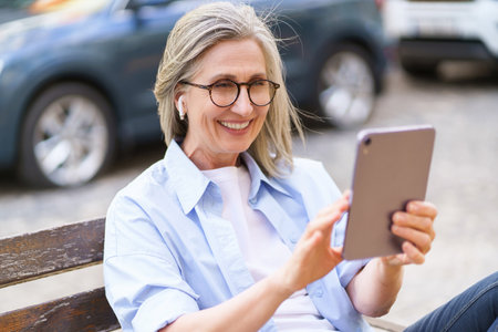 Happy and confident mature woman smiles while holding tablet PC and taking call in busy city. The urban scene and modern technology convey sense of connectivity and convenience, while the womans positive energy and independent spirit inspire success and vibrancy. High quality photoの写真素材