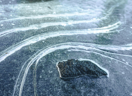 Dark, rugged rock formation standing amidst frozen landscape of icy curves and snow. Contrast of black, white, and blue colors creates dramatic and desolate scenery, conveying raw beauty and challenges of remote and untamed wilderness. High quality photoの写真素材