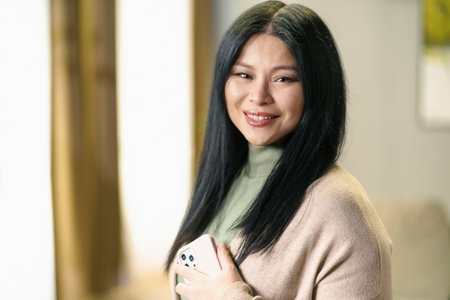 Mature and positive Asian woman with thick, luscious hair holds her smartphone in her hand, smiling with contentment and confidenced. High quality photoの写真素材