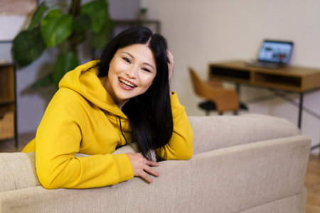 Beautiful middle-aged Asian woman is captured resting on a sofa in her home, surrounded by the comfort and coziness of her indoor space. The background reveals a desktop with a laptop, symbolizing the balance of work and leisure in her lifestyle. High quality photoの写真素材