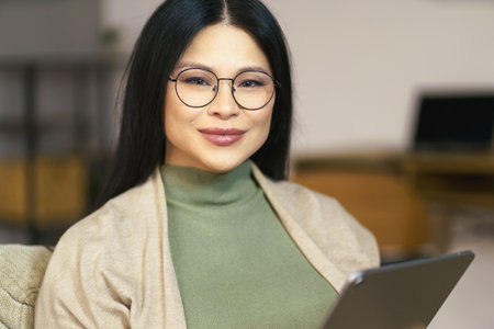 cheerful middle-aged Asian woman wearing glasses enjoys browsing internet on her tablet while sitting comfortably at home. High quality photoの写真素材
