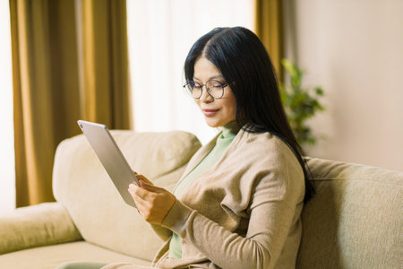 Mature Asian woman wearing glasses is deeply focused on browsing internet on her tablet while comfortably seated in the interior of her home. High quality photoの写真素材