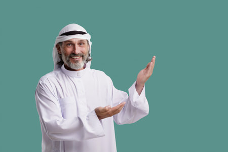 Joyful Arab man wearing Dishdasha, traditional clothing, smiles and points both palms towards copy space on green background. He is isolated and ready to add meaning to your project. High quality photoの写真素材