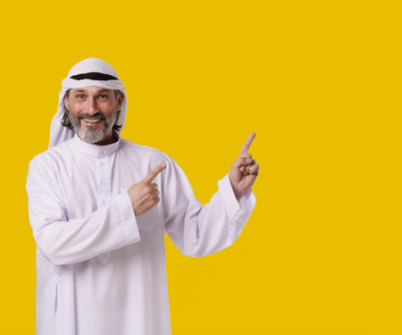 Smiling Arab man captures his cheerful and confident personalit, he points both hands towards copy space on isolated yellow background. Copy space for designers and publishers to add their text, logos. High quality photoの写真素材
