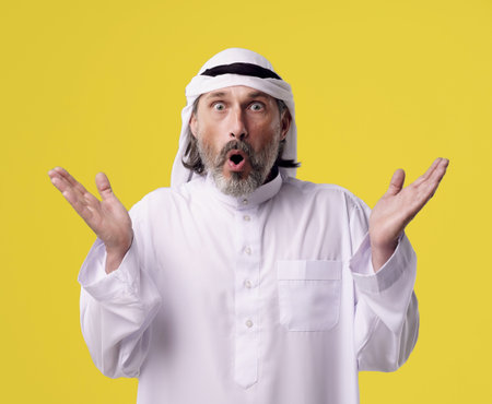 Surprised Arab man in traditional Dishdasha clothing raises both hands in the air, isolated on a bright yellow background with copy space. High quality photoの写真素材
