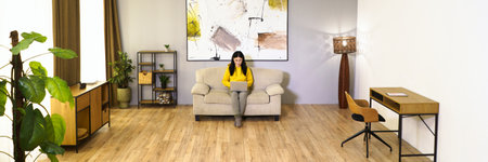 Young woman is working on laptop while sitting on couch in her comfortable living room. She is engaged in digital work, enjoying benefits of remote work and modern technology. High quality photoの写真素材