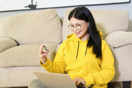 Asian woman comfortably shopping online from her home, with laptop on her lap and bank card in her hand. She is engaged in digital retail, using modern technology and the convenience of e-commerce to make purchase with her credit card. High quality photoの写真素材