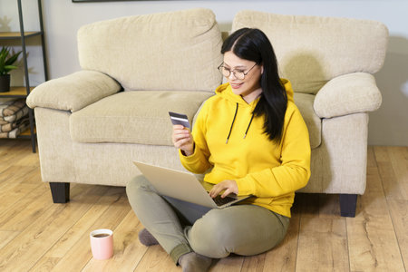 Mature Asian woman wearing glasses is sitting comfortably on floor with laptop, holding bankcard, and cup of coffee. She is engaged in digital work, enjoying moment of relaxation with her favorite beverage while working from home. High quality photoの写真素材