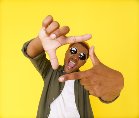 Creative young man on yellow background is seen making composition frame from his fingers, showcasing his artistic and imaginative abilities. Concept of innovation and inspiration, using hand gestures as non-verbal communication tool. High quality photoの写真素材