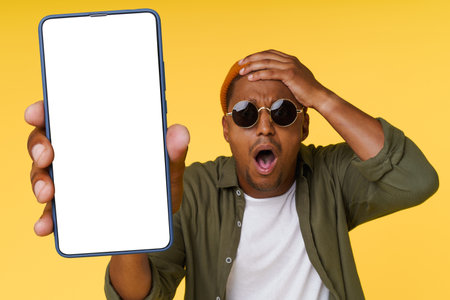 Surprised African man holds his head while showing phone with white blank screen on yellow background, concept for advertising. High quality photoの写真素材