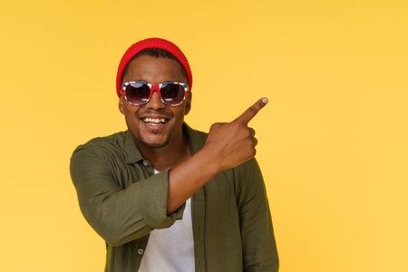 Smiling African man wearing sunglasses points to copy space on yellow background. Advertising concept. High quality photoの写真素材