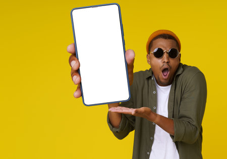 Surprised African man is holding phone with white blank screen against yellow background with copy space. The image is ideal for advertising concepts. High quality photoの写真素材