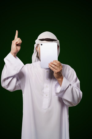 Muslim anonymous concept. Arab person holds digital tablet, face covered, index finger up, representing the concept of Islamic censorship online. Image portrays issues of privacy, security, and anonymity in the digital world. High quality photoの写真素材