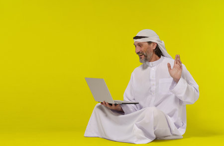 Concept online communication. Muslim person sitting on floor greets someone with their palm raised while holding a laptop in their hand. Copy space allows for easy customization and versatility and global connection through technology. High quality photoの写真素材