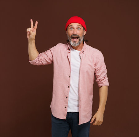 Positive European caucasian man handsome shows gesture of V victory. Cheerful man with silver beard raises his hand with two fingers V shape, indicating gesture of victory, with smile on face. High quality photoの写真素材