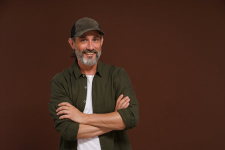 Happy Smiling man. A fisherman in a baseball cap and green shirt stands cross-legged against a brown background with copy space. High quality photoの写真素材