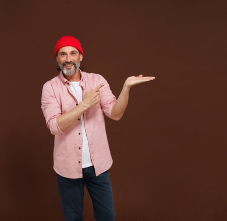 Product placement concept. Friendly bearded handsome man points his hand to copy space isolated on brown background. High quality photoの写真素材