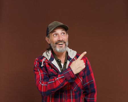Cheerful American farmer or countryman with beard smiles and points his finger to copy space for advertisement or ad, looking with delight. Happy track driver concept. High quality photoの写真素材