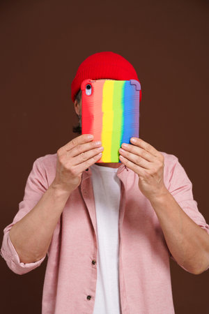 Incognito internet gay user. The man hides his sexual orientation online, covers his face with tablet in rainbow case, as symbol LGBTQ. High quality photoの写真素材
