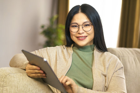 Confident middle-aged Asian woman enjoys moment of relaxation at home, browsing internet on her tablet with ease and comfort. High quality photoの写真素材