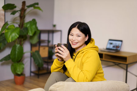 Cute Asian woman takes break from her online work at home to enjoy a soothing cup of tea. The image captures the balance of work and leisure in her comfortable indoor space, surrounded by the calm and serenity of nature. High quality photoの写真素材